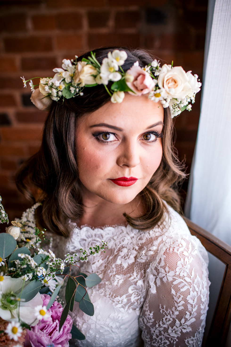 beautiful bride adorned with fresh flowers and red lipstick sitting next to the window at Curradine barns just before she gets married