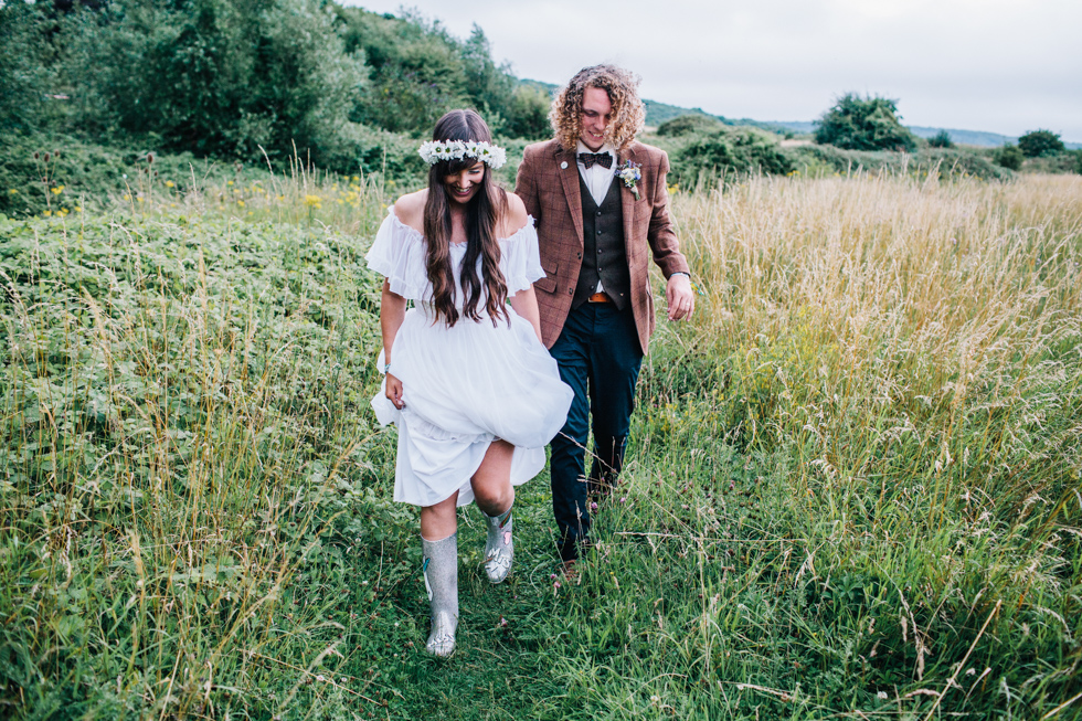 couple portrait of bohemian bride and long haired groom walking through fields at festival wedding