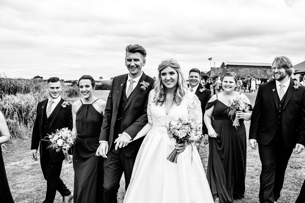 bride and groom holding hands walking and laughing with their wedding party outside 