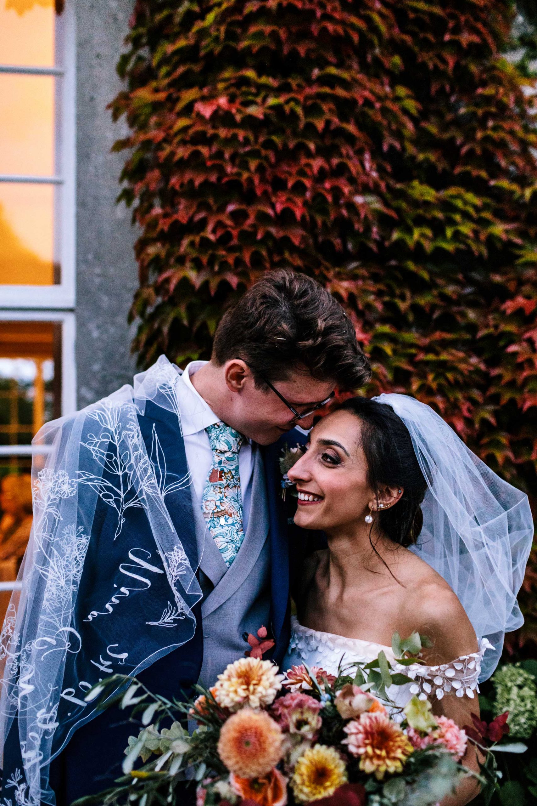 Persian bride and English groom embrace at Findon place in Surrey with autumnal flowers and personalised veil