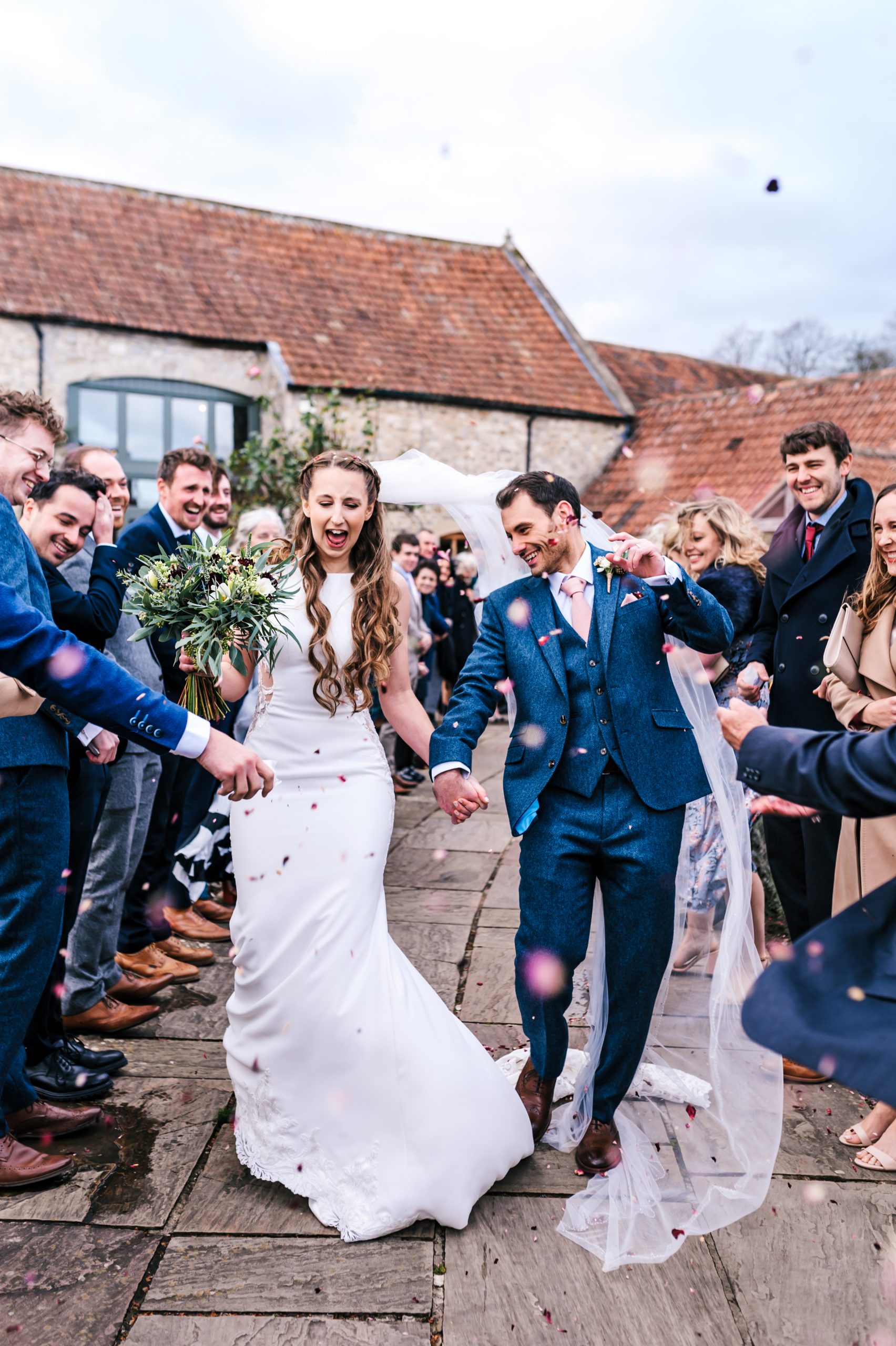 Priston Mill wedding with couple walking through confetti on a very windy day with the brides veil blowing in the wind and guests having a good time laughing.